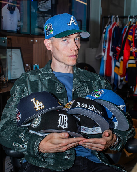 A person seated indoors holding several baseball caps, wearing a blue Atlanta Braves cap and a green patterned jacket. The hands display multiple caps with team logos and patches, including a Los Angeles Dodgers cap, a Detroit Tigers cap, and another cap with 'Los Angeles' text.
