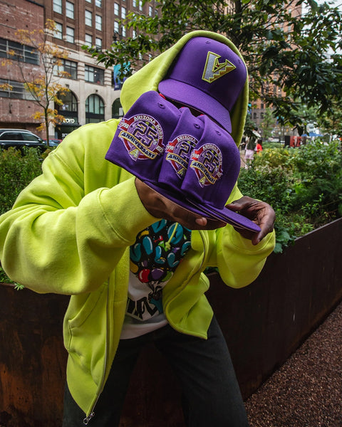 A person outdoors wearing a bright neon yellow-green hoodie and holding three purple baseball caps with a large gold 'V' logo and a '25th Anniversary' patch. The background shows a city street with trees and buildings.